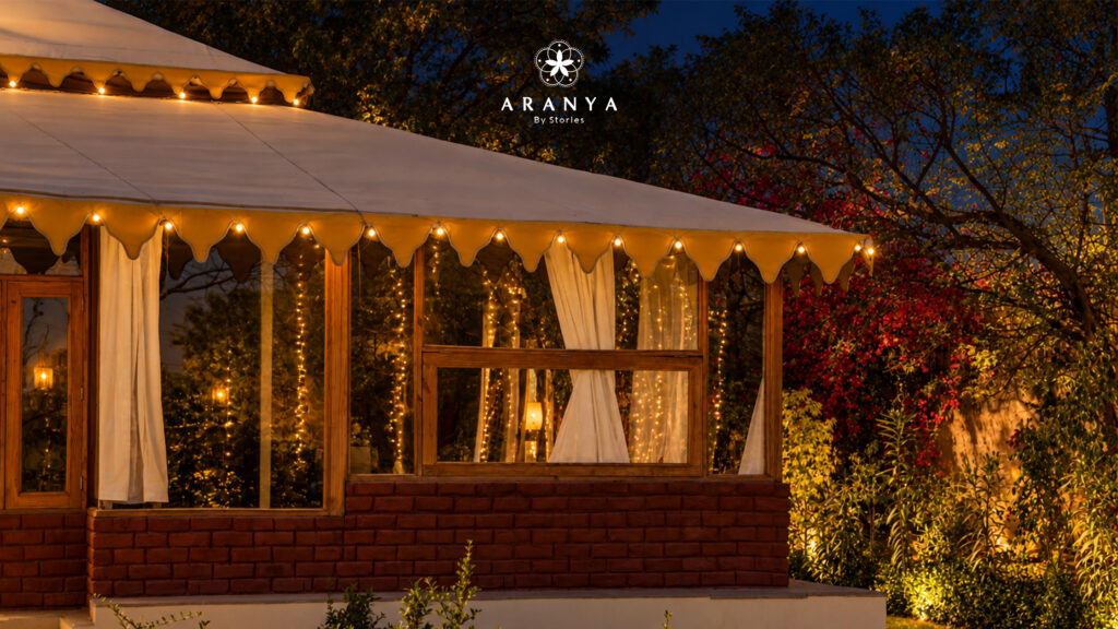 Evening view of a white canvas luxury tent with a scalloped golden valance and warm string lights, surrounded by lush trees and red bougainvillea, with the Aranya By Stories logo at the top.